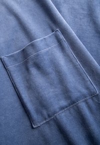 Blaue Baumwollstoff mit einem rechteckigen aufgesetzten Taschenstück, strukturierter Oberfläche und dezenten Nähten entlang der Kanten.