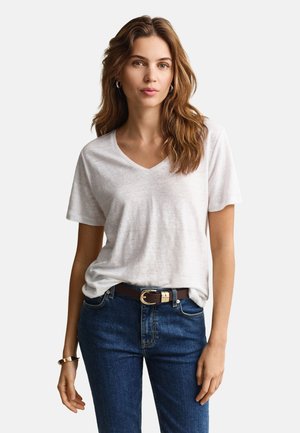 Femme aux cheveux bruns ondulés portant un t-shirt blanc à col en V, un jean bleu, une ceinture sombre, des boucles d'oreilles argentées en anneau et un bracelet sur un fond uni.