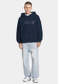 Granatowy dres z kapturem z logo 'PROSTO', przednia kieszeń kangurza i ściągacze. W połączeniu z jasnoniebieskimi jeansami o prostym kroju i białymi sneakersami.