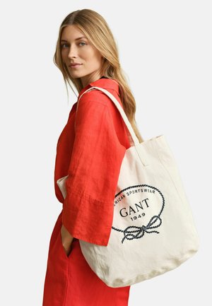 Donna bionda in abito rosso che porta una borsa tote bianca con il logo "GANT 1949" in testo nero e design a corda.