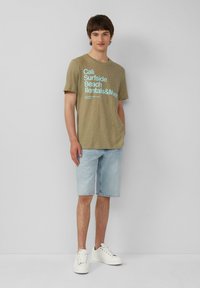 Olivgrünes T-Shirt mit blauer Schrift, kombiniert mit hellblauen Jeansshorts und weißen Sneakers. Lässig, lockere Passform. Glatte Textur.