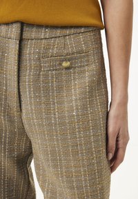 Gros plan sur un pantalon en tweed brun et gris texturé avec une poche à rabat à bouton doré, porté avec un haut moutarde et une main détendue sur le côté.