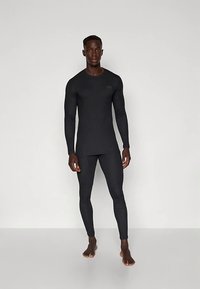 Mladi moški, bosega stoji, oblečen v oprijet črn dolg rokav in ujemajoče se pajkice na enobarvni beli podlagi.