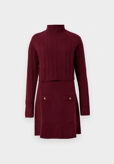 Burgundy knitted dress with a high ribbed neck, long sleeves, and a fitted skirt featuring two buttoned pockets at the front.