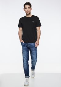 Sort bomulds T-shirt med et firkantet logo, parret med lyseblå denimjeans og hvide sneakers, sat mod en ensfarvet lys baggrund.