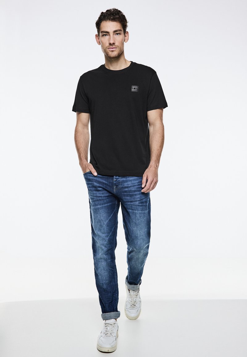 Sort bomulds T-shirt med et firkantet logo, parret med lyseblå denimjeans og hvide sneakers, sat mod en ensfarvet lys baggrund.