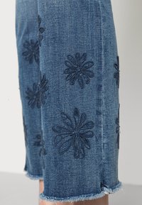 Džínové kalhoty s vyšívanými modrými květinami, třepeným lemem a slim fit. Tkanina je měkká s strukturovaným designem.