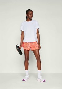 T-shirt blanc à manches courtes, shorts à motifs orange, tenant une bouteille d'eau noire, portant des chaussettes blanches et des chaussures de sport violettes claires.