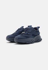 Zapatilla azul marino con parte superior de malla, lengüeta acolchada y suela de goma texturizada. Cuenta con cierre de cordones y una pestaña en el talón.