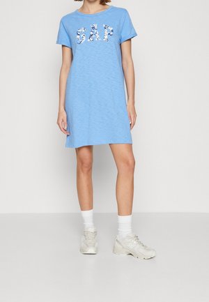 Woman wearing a light blue short-sleeve dress with "GAP" logo and white sneakers with white crew socks, standing on a gray floor against a white wall.