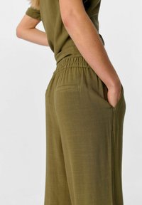 Pantalons larges vert olive avec une taille élastique, dotés de deux poches latérales et d'un tissu doux et texturé. Design minimaliste.