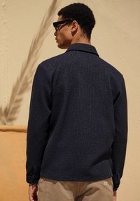 Camicia testurizzata blu navy con colletto, caratterizzata da polsini con bottoni e sottili dettagli di cucitura verticale, indossata con pantaloni khaki.