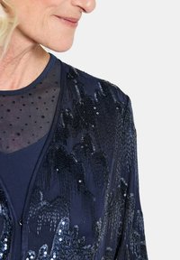 Marineblaue Paillettenjacke mit transparenter gepunkteter Mesh-Oberfläche, die ein strukturiertes Muster und eine glatte Innenschicht aufweist. Reißverschluss vorne.