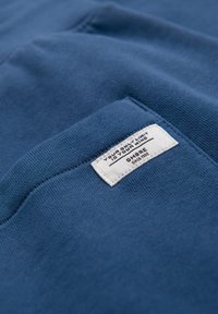 Blå bomullstyg med en texturerad yta, med en vit etikett med svart text som detaljerar varumärket och ett motiverande citat.