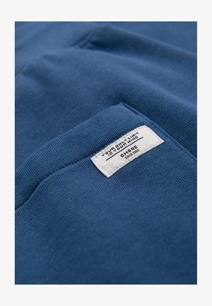 Tissu en coton bleu avec une surface texturée, présentant une étiquette blanche avec du texte noir détaillant la marque et une citation motivante.