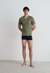 Olivgrünes kurzärmliges T-Shirt mit kleinem Logo, kombiniert mit engen schwarzen Shorts. Das Modell steht barfuß auf einem hellen Teppich.
