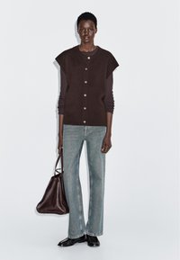 Osoba ubrana w ciemnobrązowy bezrękawnik na długim rękawie, jasnoniebieskie dżinsy, czarne buty, trzymająca dużą ciemnobrązową torbę.