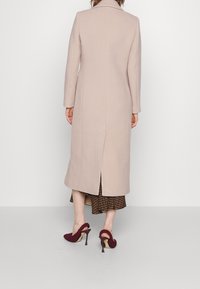 Lange beige jas met een rechte snit en een split aan de achterkant, gecombineerd met bordeauxrode, gestructureerde hoge hakken.