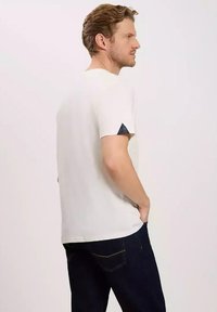 Biała bawełniana koszulka z krótkimi rękawami, z trójkątnym granatowym akcentem na lewym rękawie, zestawiona z ciemnymi dżinsami.