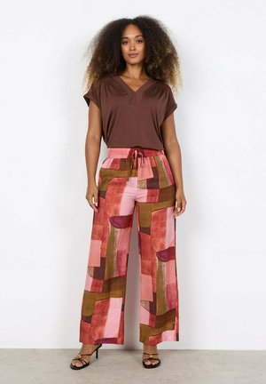 Vrouw met volumineus krullend haar, gekleed in een bruin kortemouwshirt en wijd uitlopende broek met abstracte roze, rode en bruine patronen.