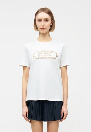 Νεαρή γυναίκα με κοντά ξανθά μαλλιά φοράει λευκό μπλουζάκι με χρυσό λογότυπο από καρφιά και μαύρη πλισέ φούστα, στέκεται μπροστά σε απλό φόντο.