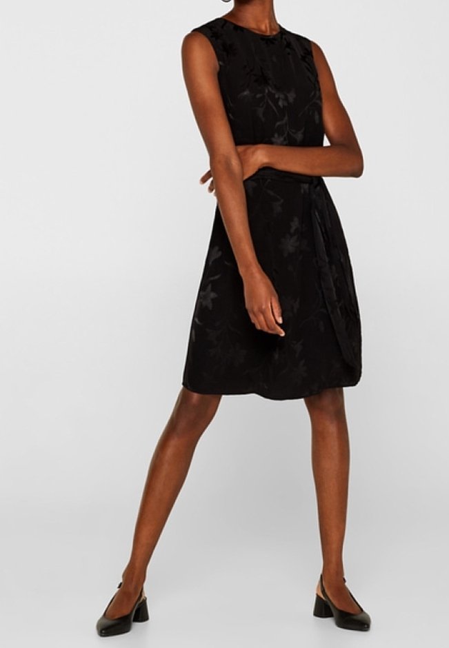 Femme portant une robe noire sans manches longueur genou avec un motif floral subtil et des chaussures noires à talons blocs, debout, bras croisés.