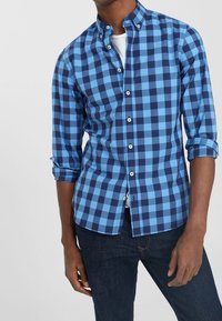 Niebieska koszula w kratę z kołnierzykiem zapinanym na guziki, z jasnoniebieskimi i ciemnoniebieskimi kwadratami. Długie rękawy podwinięte przy mankietach. Nosi się ją na białej koszuli.