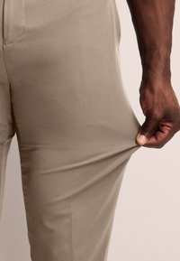 Pantalones beige hechos de una tela elástica, con una textura suave. Una mano tira de la tela para mostrar su flexibilidad y ajuste.