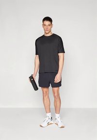 Musta lühikeste varrukatega sportlik t särk, mida kannavad tumesinised lühikesed püksid. Modell hoiab musta veepudelit; jalas valged ja hallid spordijalanõud.