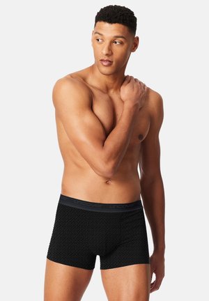 Sorte boxer brief med et mønstret tekstur og en grå elastisk linning med ordet "uncover" trykt i sort. Modellen poserer afslappet.