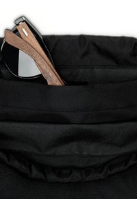 Occhiali da sole neri con aste in legno parzialmente visibili nella tasca esterna di una borsa nera in tessuto strutturato.