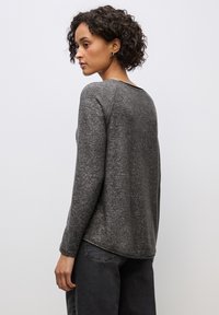 Maglione grigio a maniche lunghe con una finitura testurizzata, scollatura rotonda e orlo posteriore leggermente più lungo. Indossato con jeans scuri e larghi.