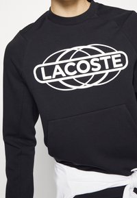 Černá mikina s bílým kruhovým logem s nápisem "LACOSTE." Má kulatý výstřih, dlouhé rukávy a přední klokaní kapsu.