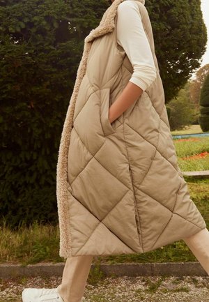 Veste longue matelassée beige avec un col en fourrure douce et doublé, dotée de poches latérales et d'une texture lisse, portée sur un haut blanc et un pantalon clair.