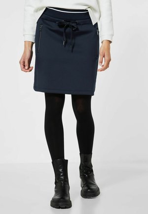Jupe bleu marine avec une taille à cordon de serrage, poches latérales zippées et une texture lisse. Associée à des leggings noirs et des bottines.