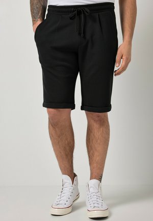 Mand iført sorte shorts med op-rullet kant og snøre i taljen samt hvide høje sneakers, står med afslappede hænder ned langs siderne.