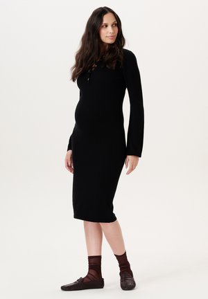 Zwangere vrouw met lang haar, gekleed in een nauwsluitende zwarte jurk met lange mouwen, bruine doorschijnende sokken en bruine loafers, staand en zijwaarts kijkend.