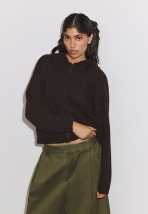 Jeune femme aux longs cheveux foncés portant un sweat à capuche noir zippé et un pantalon taille haute vert olive, debout devant un fond blanc.