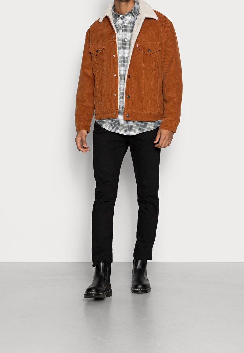 Homme portant une veste en velours côtelé couleur rouille avec col sherpa blanc, chemise à carreaux grise, pantalon noir et bottes en cuir noires, debout contre un mur blanc.