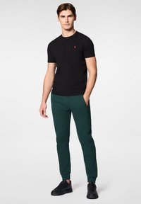 Czarna koszulka z okrągłym dekoltem, czerwona aplikacja na piersi; ciemnozielone dopasowane dresy z kieszeniami na zamek; czarne sneakersy z teksturowaną powierzchnią.