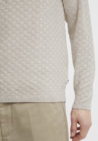 Beige textured sweater with a checkerboard pattern, ribbed cuffs and hem. Worn over light-colored trousers, showcasing the fabric's knitted detail.