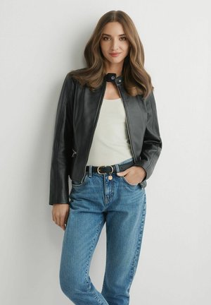 Kobieta z rozpuszczonymi brązowymi włosami, ubrana w czarną skórzaną kurtkę, biały top, niebieskie dżinsy i czarny pasek, stojąca przy białej ścianie.