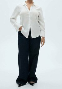 Chemise en lin blanc avec col, manches longues et coupe décontractée, associée à un jean bleu marine à jambes larges et des chaussures noires pointues.