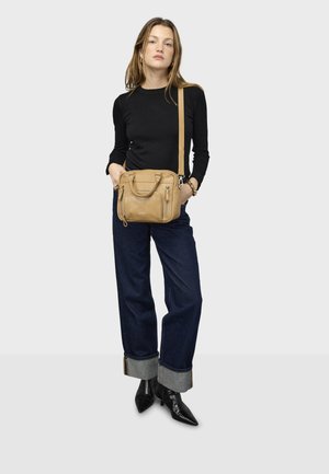 Bruine leren schoudertas met ritssluitingen, rechthoekige vorm en een afneembare band. Het model draagt een zwarte top, wijde blauwe jeans en zwarte laarzen.