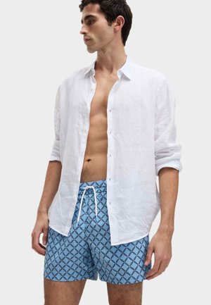 Weißes Leinen-Hemd mit Knopfleiste, über blauen Badehosen mit einem geometrischen Muster und dunklen Konturen getragen. Das Model steht mit einem neutralen Ausdruck.
