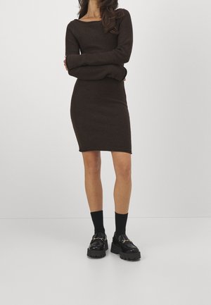 Woman wearing a fitted long-sleeve dark brown dress, black socks, and chunky black loafers, standing with arms crossed against a plain background.