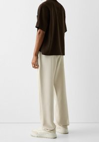 Hnědá košile s krátkým rukávem v kombinaci s krémovými širokými kalhotami a světlými teniskami. Košile má volný střih a kalhoty hladkou texturu.