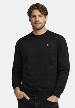 Mann mit welligem braunem Haar, der einen schwarzen Sweatshirt mit Rundhalsausschnitt und einem kleinen roten Logo auf der linken Brust trägt, Hände in den Taschen.