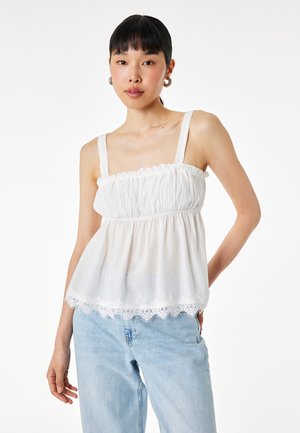 Jeune femme portant un haut blanc sans manches froncé avec un bord en dentelle et un jean bleu clair, debout devant un fond blanc uni.