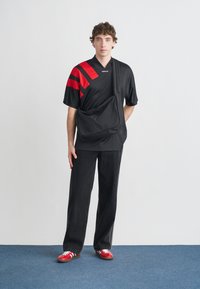 Čierne tričko s červenými diagonálnymi pruhmi, bočná taška cez rameno, doplnené voľnými čiernymi džínsami a červenými teniskami s bielymi detailmi.